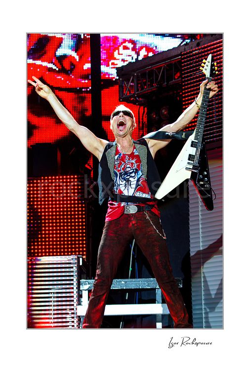 Vertical color image of Rudolf Schenker of Scorpions performing live with raised arms, white guitar, and vivid red stage lights