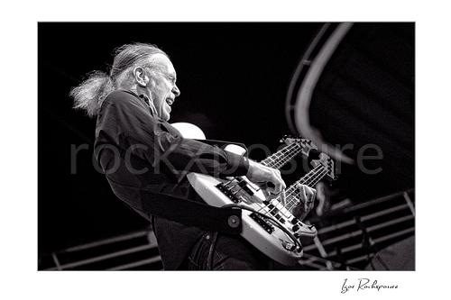 Horizontal black and white image of Billy Sheehan playing his double-neck bass in an energetic side-stage performance shot