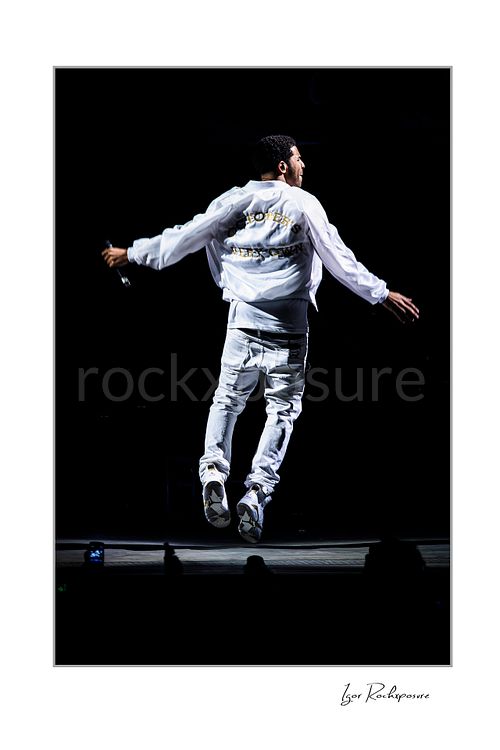 Vertical color image of Drake performing live on stage from behind in white outfit mid-jump against a black background