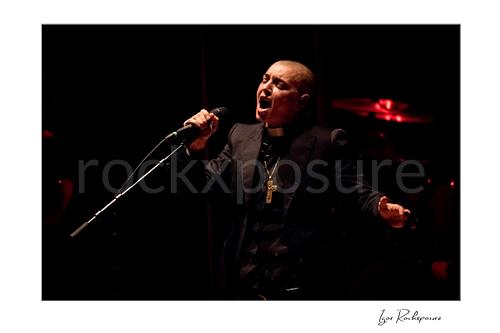 Horizontal color image of Sinéad O’Connor singing into a microphone on stage in a dramatic low-light portrait
