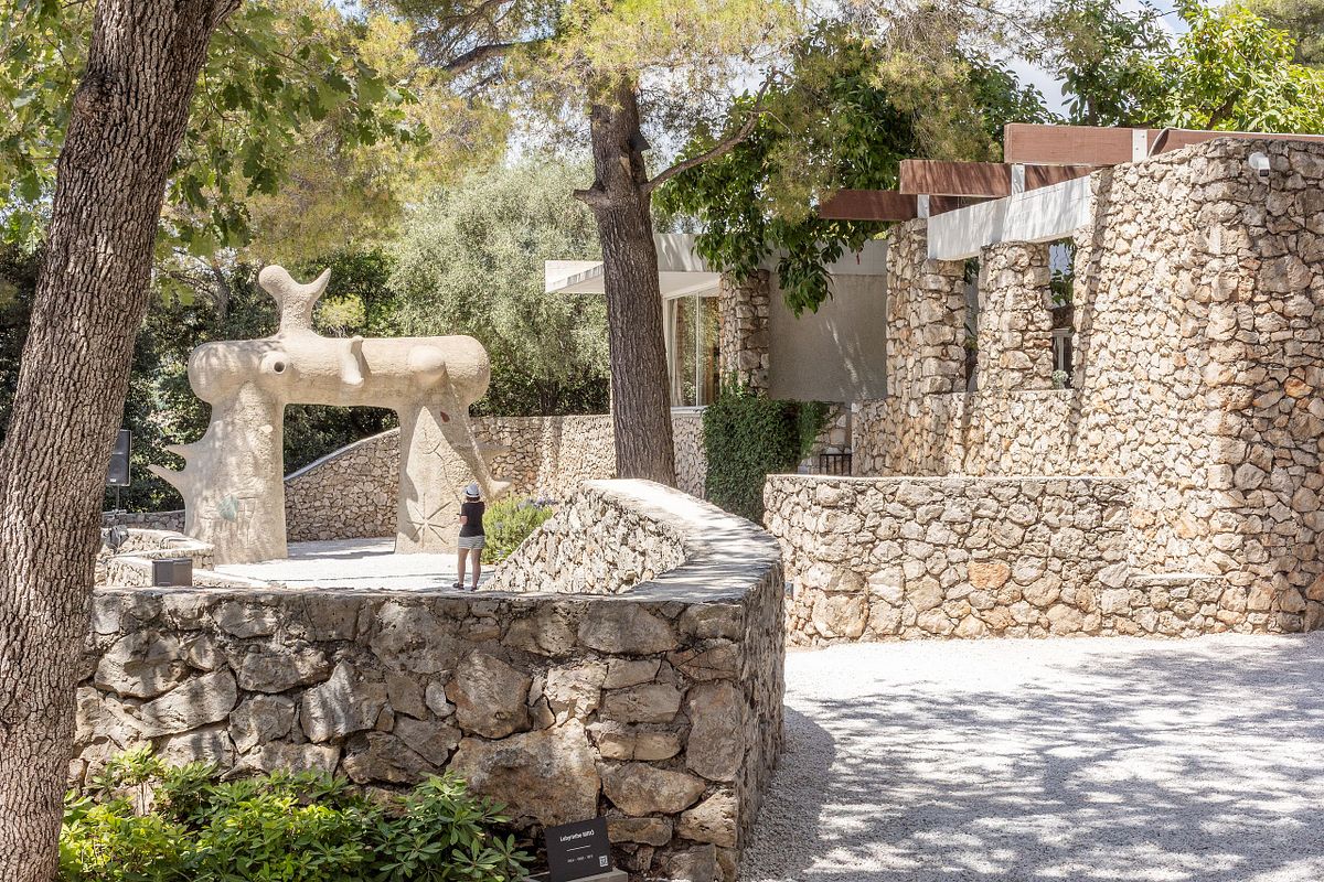Bes&ouml;kare i skulpturpark med stenmur och stor abstrakt skulptur i soligt dagsljus.
