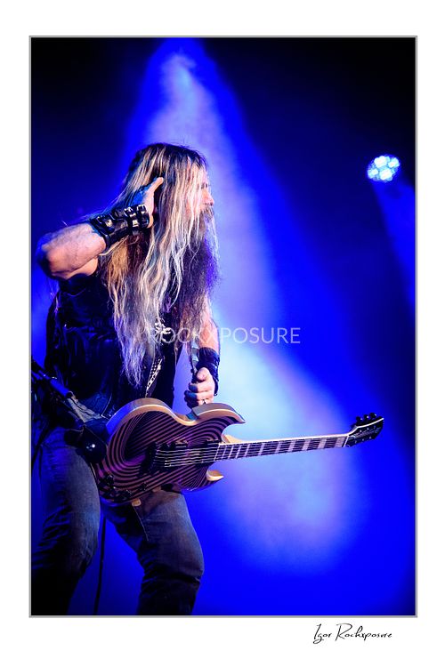 Vertical shot of Zakk Wylde playing guitar on stage, wearing a denim vest, looking to the side under an intense blue spotlight and atmospheric stage fog.