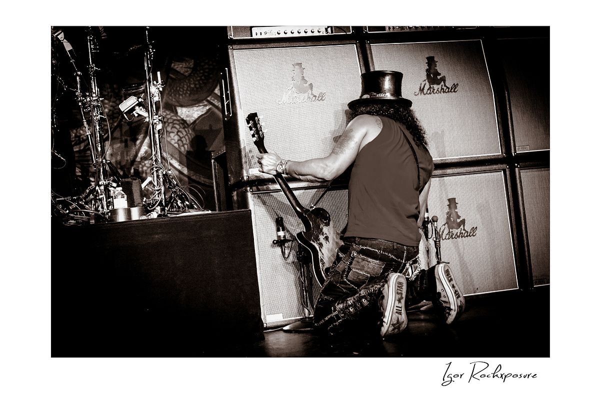 Horizontal concert photography of Slash sitting with his back to the audience holding his electric guitar toward a wall of amplifiers in black and white photography under stage lighting