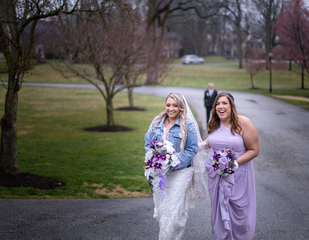 bride and her maid-of-honor is walking on wet pavement to the ceremony site. she is wearing a jeans jacket and her flower bouquet