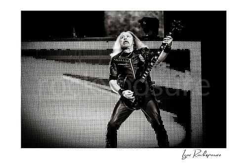 Horizontal black and white image of Glenn Tipton of Judas Priest performing live on stage, playing electric guitar in a wide stance against a bright LED screen backdrop