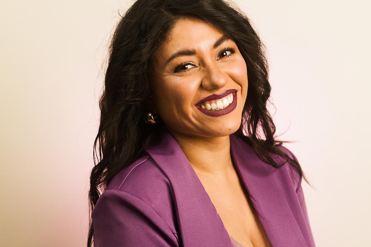 Retrato de una mujer sonriente, con saco morado, con aspecto amable y emprendedor.