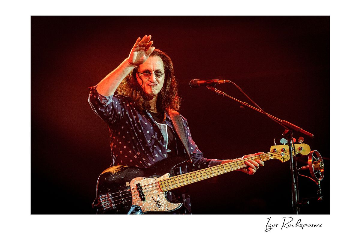 Horizontal concert photography of Geddy Lee playing bass guitar beside a microphone under red stage lighting