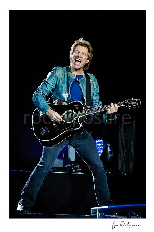 Vertical color image of Jon Bon Jovi performing live with a black acoustic guitar, smiling broadly on stage with blue concert lights behind him