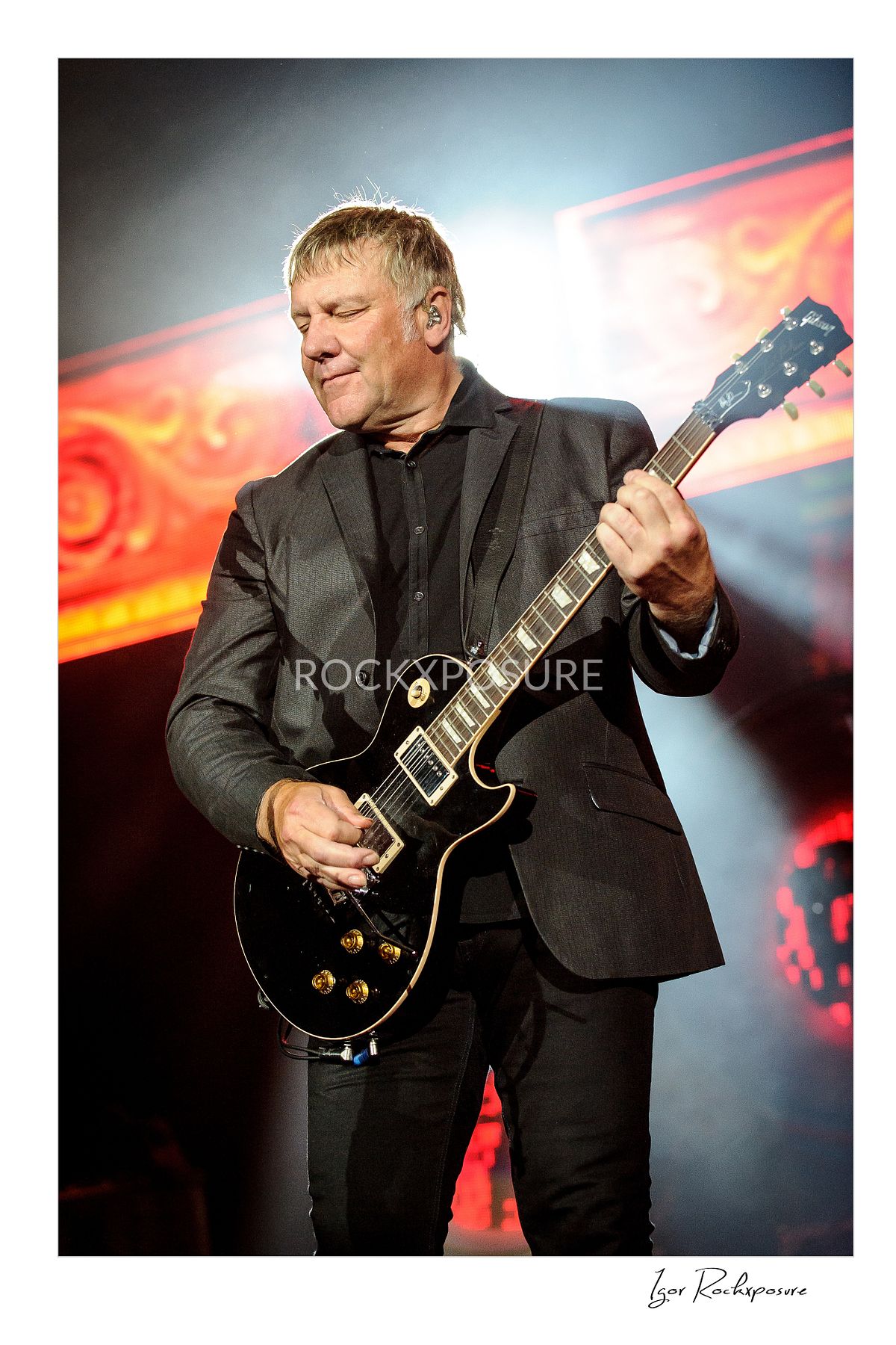 Vertical concert photography of Alex Lifeson playing electric guitar under colorful stage lighting