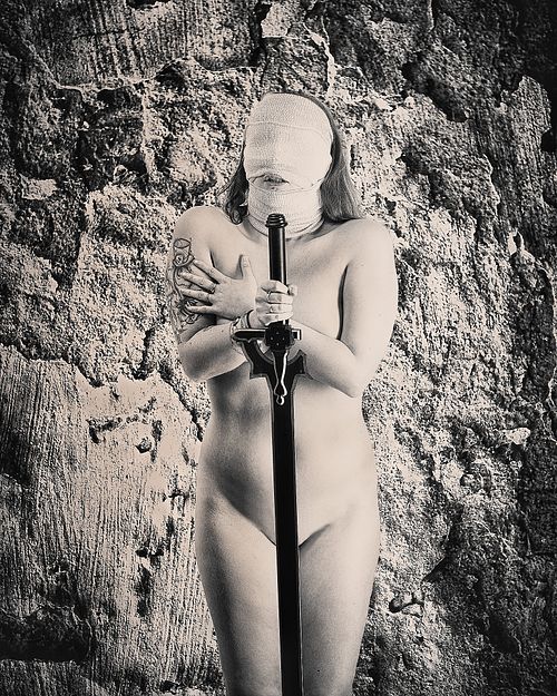 photo tarot L’Épreuve, femme dos au mur yeux bandés tenant une épée baissée, dans une posture de vulnérabilité et défi