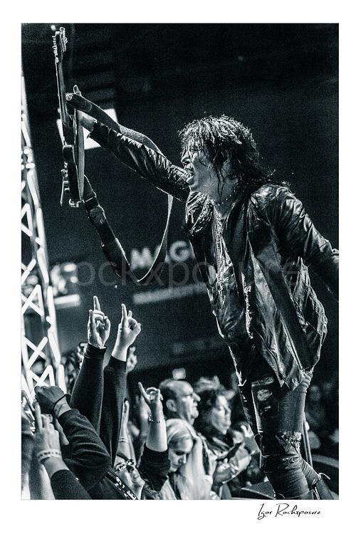 Vertical black and white image of Tom Keifer performing live at the edge of the stage, raising his black guitar overhead toward the crowd in a fierce concert moment