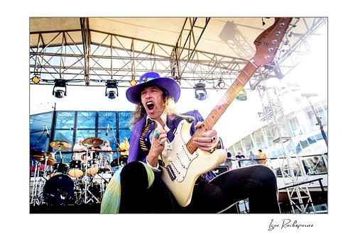 Horizontal color image of Randy Hansen playing a white guitar on an outdoor ship deck stage while wearing a purple hat and jacket