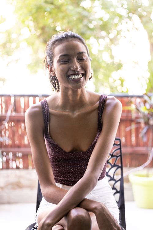 A portrait of an Indian woman, smiling and wearing a purple top, sitting on a chair on a balcony with her arms placed on her lap.
