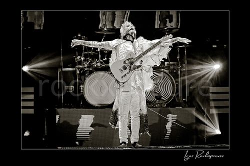 Horizontal black and white image of John 5 performing live on stage with an electric guitar, white stage outfit, outstretched arms, and bright backlights