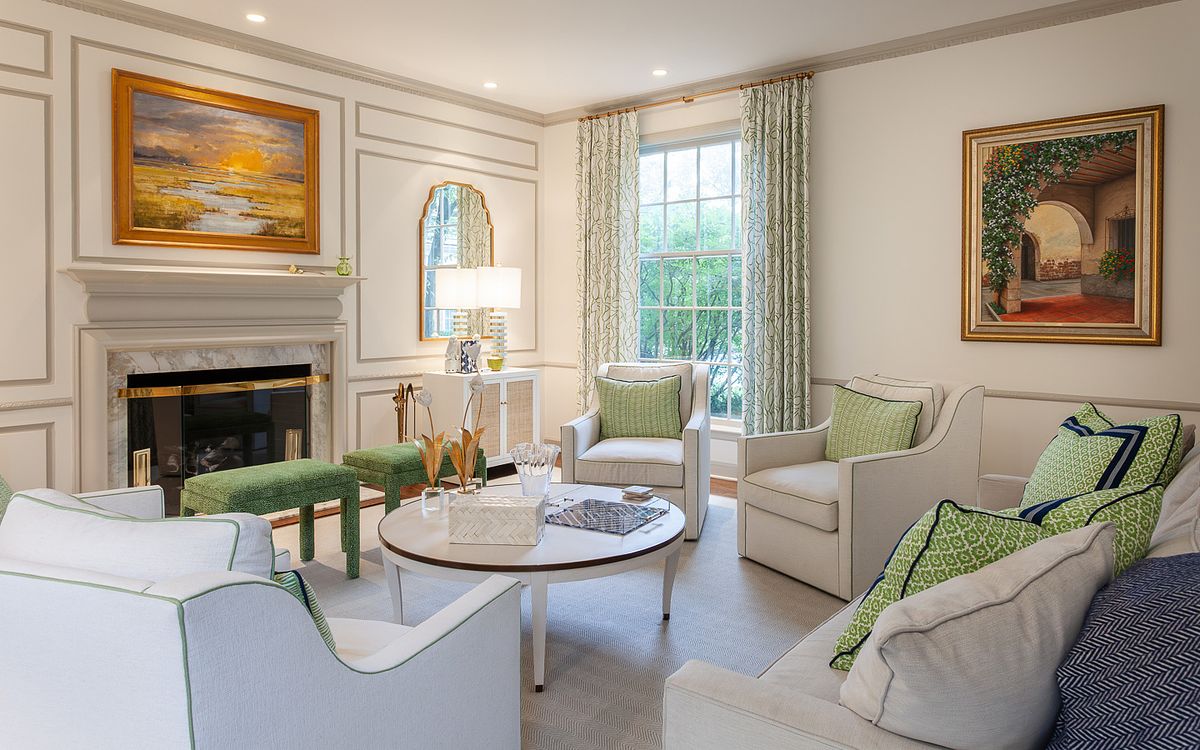 Bright and sophisticated living room featuring white upholstered armchairs with green piping, a marble fireplace, and classic gold framed landscape art on wainscoted walls