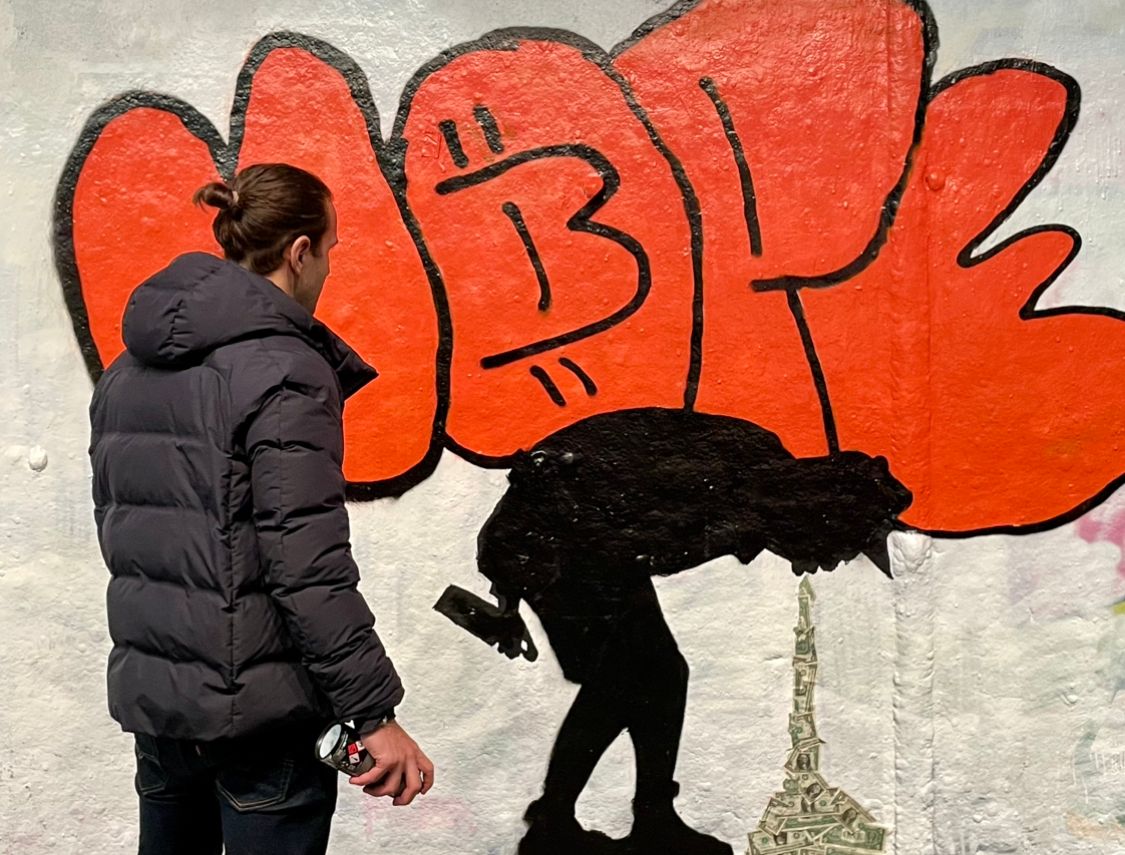 Street art mural by Street Cyber in London depicting a graffiti tag "HO₿E" in orange. Beneath it, a dark stencil figure appears to be expelling or 'vomiting' a stream of real dollar bills that form a tower, subtly referencing structures like the Eiffel Tower, symbolizing fiat money. This powerful piece critiques the inflationary nature of traditional currency, transforming it into a tangible visual of waste and the hope found in Bitcoin