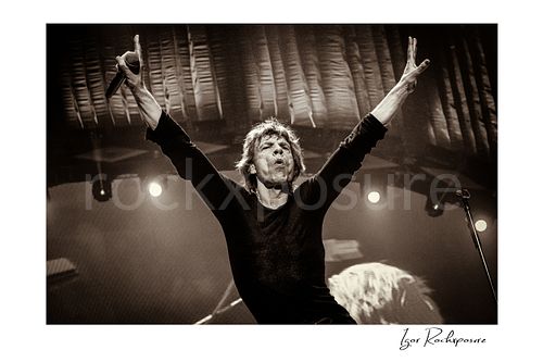 Horizontal black and white image of Mick Jagger of The Rolling Stones raising both arms on stage during a live performance