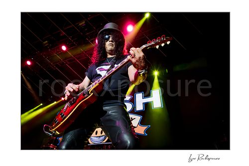 Horizontal concert photography of Slash playing electric guitar wearing his signature top hat under colorful stage lighting