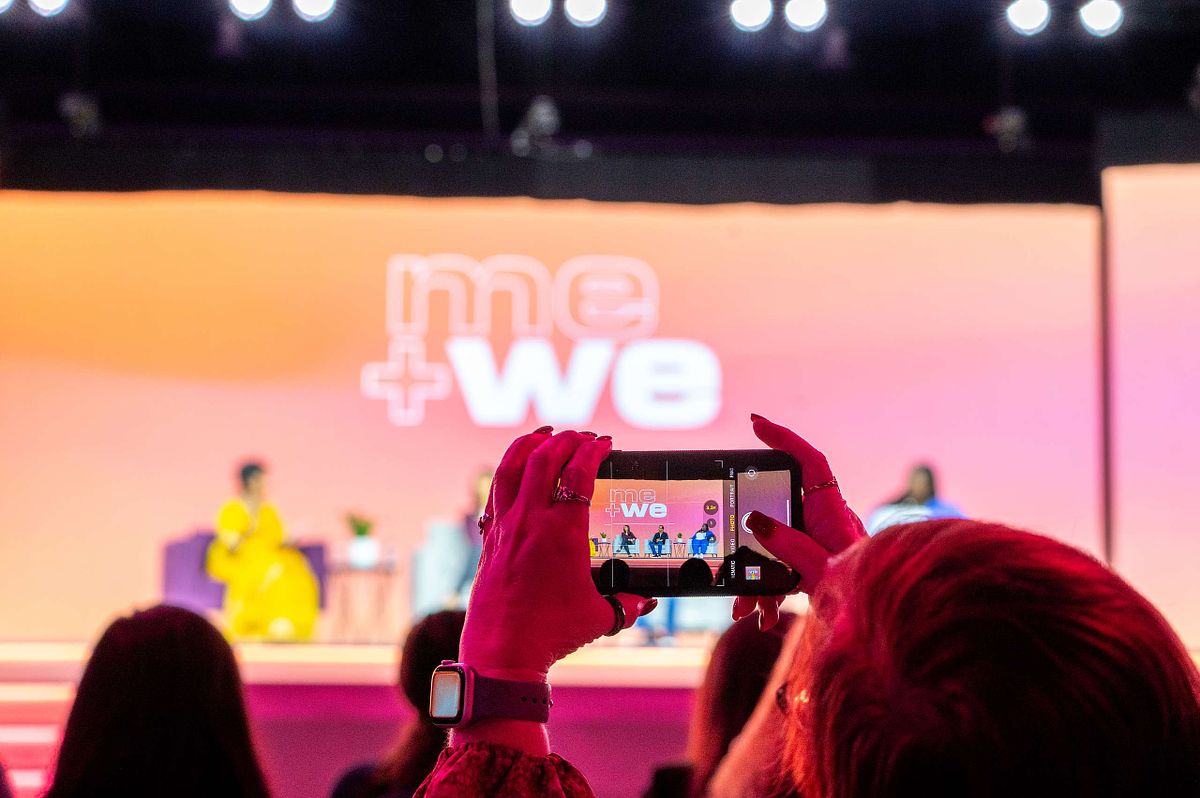 Audience member records &ldquo;me + we&rdquo; panel at a corporate event using her smartphone, capturing content for social media.