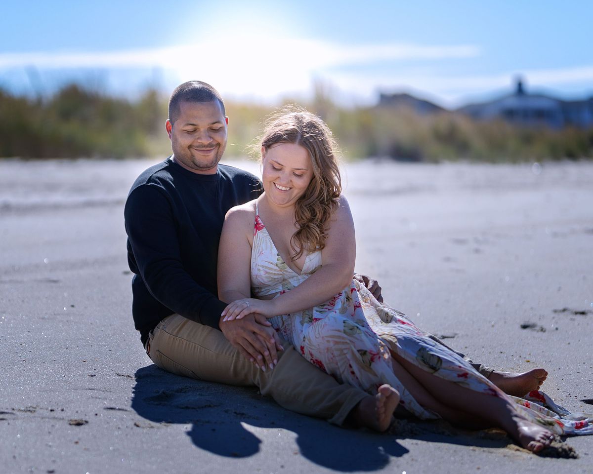 couple sitting on the beach in cape henlopen near lewes delaware during their engagement session looking at the engagement ring